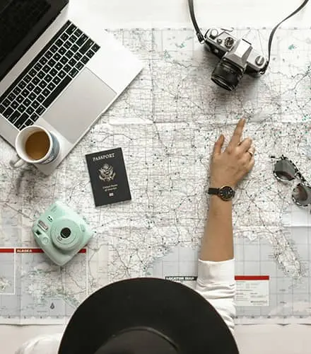 Woman pointing to a spot on a map spread out on a table. There is a laptop, coffee cup, camera, passport and sunglasses on top of the map (certified professional career coach)