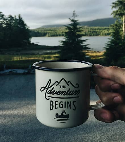 Person sitting near a lake with some pine trees holding a mug that has 'the Adventure Begins' written on it (CPCC certification)